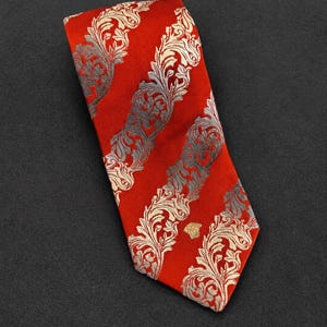 May include: A vibrant red necktie featuring diagonal stripes of intricate, light-colored, ornate scrollwork patterns. The fabric has a subtle sheen, and a small, gold-toned Medusa head emblem is positioned near the pointed bottom edge of the tie.