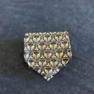 Peut inclure: Une cravate présentant un motif répétitif de formes dorées rappelant des feuilles et de petits accents triangulaires blancs et bleus sur un fond gris foncé texturé. Le tissu présente un léger éclat et une extrémité inférieure triangulaire.