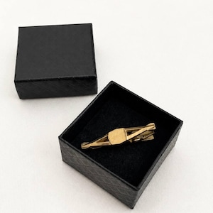 May include: A gold-toned metal tie clip with a geometric, angular design resting inside a square black gift box with a textured, quilted pattern on the exterior. The lid of the box is placed beside the open base, revealing a black velvet interior.