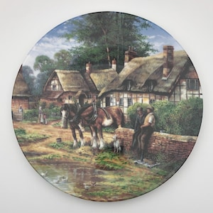 May include: Circular decorative plate with a rural scene depicting thatched cottages, horses, and figures by a pond. The artwork showcases a detailed, colourful illustration of a countryside landscape. The plate has a glossy finish and a white border.