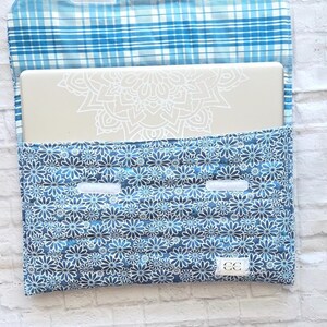 May include: A quilted fabric laptop sleeve featuring a blue and white plaid interior flap and a dark blue exterior with a repeating white daisy floral pattern. The sleeve includes two white hook-and-loop fasteners and a small white CC brand tag.
