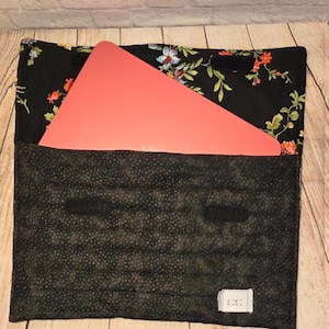 May include: A quilted laptop sleeve featuring a black fabric with a colorful floral pattern on the flap and a black fabric with small white dots on the main body. A coral-colored laptop is partially inserted, and a white tag with CC is attached.