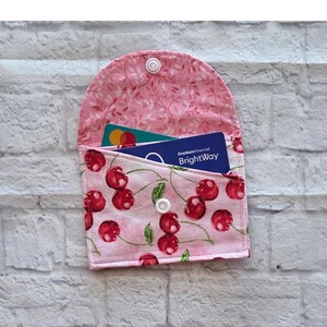 May include: A pink fabric card holder with a cherry pattern, open to reveal credit cards. The interior lining features a pink floral design. The card holder is secured with a white snap closure. The text "OneMain Financial BrightWay" is visible on one of the cards.