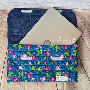 May include: A fabric laptop sleeve featuring a vibrant pattern of pink flamingos and green palm leaves on a dark blue background. The sleeve has a flap closure with two white hook-and-loop fasteners and a small white rectangular brand label.