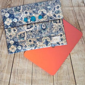 May include: A fabric laptop sleeve featuring a blue, beige, and white floral damask pattern with three teal buttons on the flap. A small white rectangular label with the letters CC is stitched on the front. The sleeve rests atop a bright orange laptop.