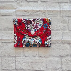 May include: A rectangular fabric pouch featuring a vibrant red background with a multi-colored pattern of stylized sugar skulls, intricate floral motifs, and swirling decorative elements. The pouch has a flap closure secured by a white snap button.