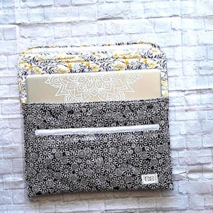 May include: A rectangular laptop sleeve featuring a black fabric exterior with a white daisy floral pattern and a flap closure lined with a yellow and gray leaf print. A silver laptop with a white mandala decal is partially inserted into the sleeve.