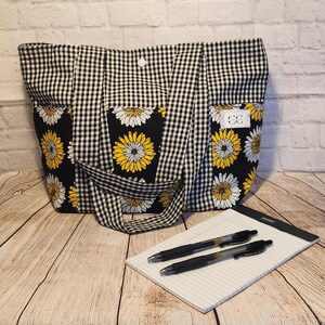 May include: A black and white gingham tote bag with pockets featuring a yellow and white daisy pattern. The bag has a button closure and handles. Two black pens and a notepad are in front of the bag.