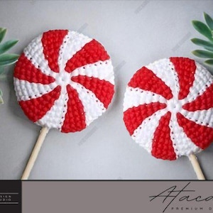 May include: Two handmade, red and white striped lollipops on wooden sticks. The lollipops are crocheted with a spiral design. The background includes a succulent plant and the text "HAZ Design Studio" and "Atacan Premium Designs".