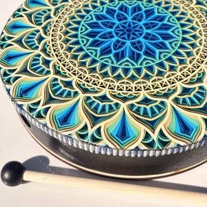 May include: A circular frame drum featuring a vibrant mandala pattern in shades of blue, teal, and cream on the drumhead. The drum has a black side frame with a textured silver trim along the top edge and includes a wooden mallet with a black ball tip.