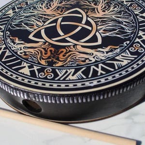 May include: A round black frame drum featuring a central triquetra symbol intertwined with a tree design in shades of cream and tan. The outer border displays geometric patterns and symbols. A plain wooden mallet rests beside the drum.