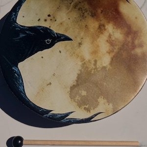 May include: A round frame drum featuring a printed graphic of a black crow flying across a large, textured, mottled beige and brown moon. The drum is accompanied by a wooden mallet with a small, round, black head for striking the surface.