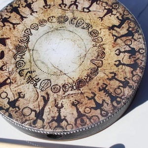 May include: A round frame drum with a textured, beige, and brown surface featuring black printed illustrations of stylized human figures and circular symbols arranged in concentric rings. A wooden mallet with a black tip lies nearby.
