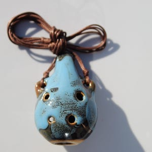 May include: A pear-shaped ceramic ocarina pendant featuring a glossy light blue glaze with dark brown speckled accents and six finger holes. The instrument is suspended from a brown cord tied in a knot at the top for wearing as a necklace.