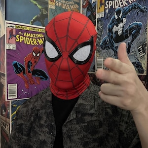 Puede incluir: Una m&aacute;scara roja de Spider-Man con telara&ntilde;as negras y malla blanca para los ojos. La m&aacute;scara cubre toda la cabeza y el cuello. En el fondo se ven portadas de c&oacute;mics de Spider-Man.