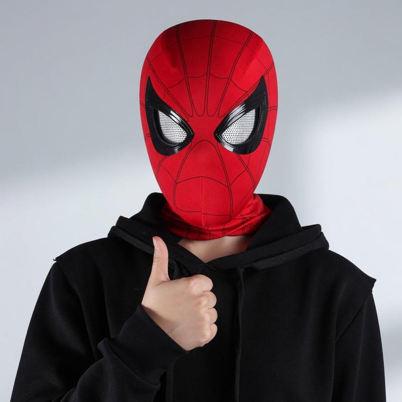 Puede incluir: Una m&aacute;scara roja de Spider-Man con l&iacute;neas negras de telara&ntilde;a y grandes aberturas para los ojos con bordes negros. La m&aacute;scara cubre toda la cabeza y el cuello. La persona lleva una sudadera con capucha negra y levanta el pulgar.