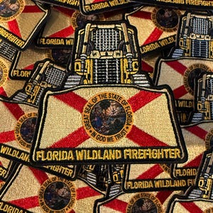May include: A collection of embroidered patches featuring a yellow bulldozer silhouette above the Florida state flag design. Each patch displays the Great Seal of the State of Florida in the center and the text Florida Wildland Firefighter in gold.