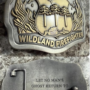 May include: A rectangular metal belt buckle with a silver finish and gold accents. The top of the buckle reads "FLORIDA WILDLAND FIREFIGHTER" with a shield design featuring a helicopter, trees, and a bulldozer. The back of the buckle has the text: "LET NO MAN'S GHOST RETURN TO SAY HIS TRAINING LET HIM DOWN."