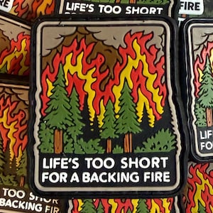 May include: A rectangular patch featuring a forest fire scene with green trees and flames in red, yellow, and orange. The text "LIFE'S TOO SHORT FOR A BACKING FIRE" is displayed in white.