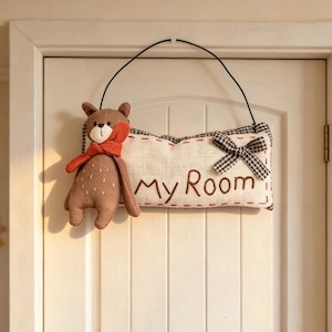 Peut inclure: Une plaque de porte rectangulaire en tissu avec le texte My Room brodé en marron. Elle est ornée d'un petit ours en peluche marron portant une écharpe orange et un nœud à carreaux noir et blanc. Elle est suspendue par un fil métallique noir.