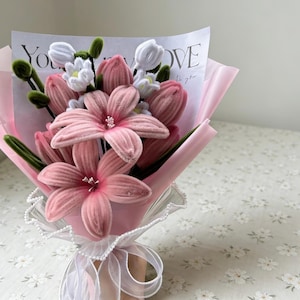 May include: A bouquet of pink fabric flowers, including lilies and tulips, wrapped in pink paper and tied with a white ribbon. The bouquet includes a card with the words "You LOVE to you".