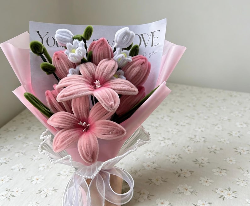 May include: A bouquet of pink fabric flowers, including lilies and tulips, wrapped in pink paper and tied with a white ribbon. The bouquet includes a card with the words "You LOVE to you".