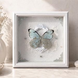 Puede incluir: Un marco cuadrado blanco tipo caja con una mariposa azul claro con detalles de purpurina plateada. La mariposa está centrada sobre un pequeño marco ovalado blanco en un fondo blanco texturizado, con adornos plateados en las esquinas.
