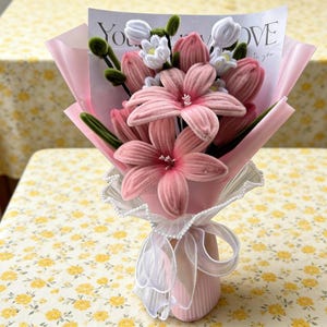 Puede incluir: Un ramo de flores rosas de tela, que incluye lirios y tulipanes, envuelto en papel rosa. El ramo está atado con una cinta blanca y encaje. Se ve una tarjeta con las palabras "You LOVE".