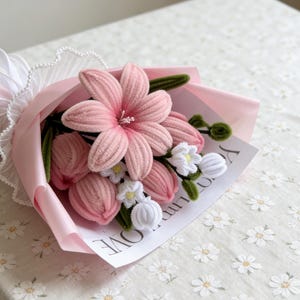 May include: A handmade bouquet of pink fabric flowers, including lilies and tulips, wrapped in pink paper and tied with a white ribbon. The bouquet includes a small card with the word "LOVE" visible.