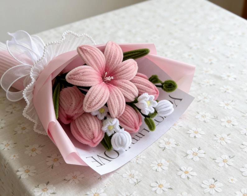 May include: A handmade bouquet of pink fabric flowers, including lilies and tulips, wrapped in pink paper and tied with a white ribbon. The bouquet includes a small card with the word "LOVE" visible.