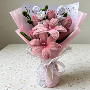 May include: A bouquet of pink fabric flowers, including lilies and tulips, wrapped in pink paper. The bouquet is tied with a white ribbon and lace, and a card with the word "LOVE" is visible.
