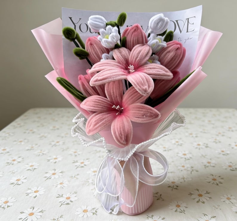 May include: A bouquet of pink fabric flowers, including lilies and tulips, wrapped in pink paper. The bouquet is tied with a white ribbon and lace, and a card with the word "LOVE" is visible.