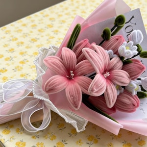 May include: A bouquet of pink fabric flowers, including large blooms and smaller white flowers, wrapped in pink paper and tied with a white ribbon. The bouquet includes a card with the word "LOVE" printed on it.