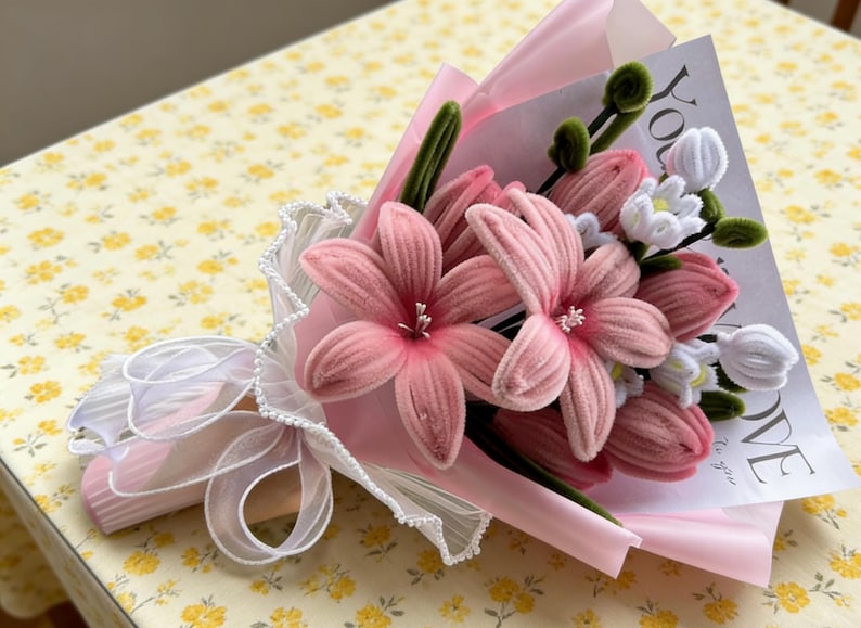 May include: A bouquet of pink fabric flowers, including large blooms and smaller white flowers, wrapped in pink paper and tied with a white ribbon. The bouquet includes a card with the word "LOVE" printed on it.