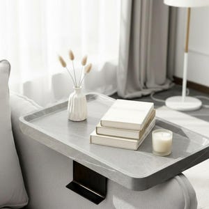 Puede incluir: Una mesa gris para el brazo del sofá con un soporte de metal negro. Tiene una pila de libros, un pequeño jarrón blanco con flores secas y una vela encendida. La escena tiene lugar en una habitación luminosa.