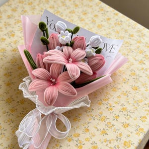 May include: A bouquet of pink fabric flowers, including lilies and tulips, wrapped in pink paper and tied with a white ribbon. A card with the word "LOVE" is visible. The bouquet is on a yellow floral patterned surface.