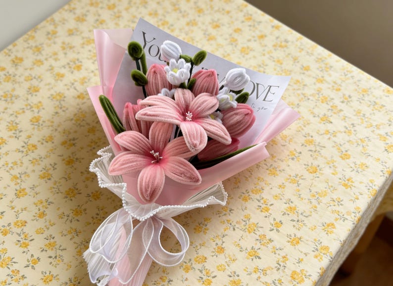 May include: A bouquet of pink fabric flowers, including lilies and tulips, wrapped in pink paper and tied with a white ribbon. A card with the word "LOVE" is visible. The bouquet is on a yellow floral patterned surface.
