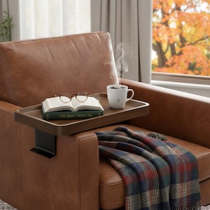 May include: A brown leather armchair with a wooden tray holding a book, glasses, and a white mug of coffee. A plaid blanket rests on the chair. The tray is attached to the armrest with a black metal bracket. The book is titled "The Quiet Scholar."