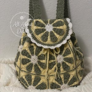 May include: A handmade crochet backpack in shades of olive green, cream, and pale yellow. The bag features a floral pattern and a drawstring closure. The flap is adorned with a white scalloped edge. The straps and body of the bag are visible.