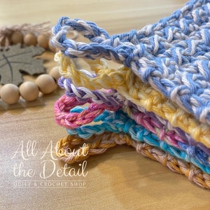May include: A stack of colorful crocheted dishcloths in blue, yellow, purple, pink, turquoise, and beige. The dishcloths are made with a textured stitch and have a loop for hanging. The text "All About the Detail" is visible in the bottom left corner of the image.