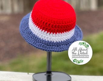 May include: A red, white, and blue crocheted bucket hat. The hat is displayed on a black stand. The text "All About the Detail Quilt & Crochet Shop" is visible in the image.