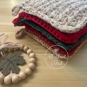 May include: A stack of handmade crochet pot holders in various colors: beige, red, dark green, and tan. Each pot holder has a loop for hanging. A wooden bead garland with a felt leaf is in the foreground.
