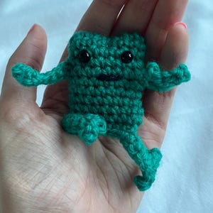 May include: A handmade, green crochet frog with black safety eyes and a smiling mouth. The frog has outstretched arms and one leg extended, giving the appearance of movement. The frog is a small, whimsical toy.