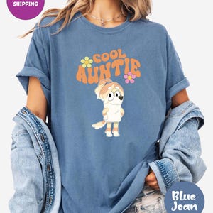 May include: A blue t-shirt with the words "COOL AUNTIE" in orange and yellow, with a cartoon character illustration. The shirt has rolled-up sleeves and is paired with a denim jacket.