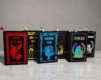 May include: Six black gift bags, shaped like books, with vibrant illustrations and titles such as "Red Rising" and "Dark Age." Each bag features a ribbon handle. The bags are arranged on a marble surface.