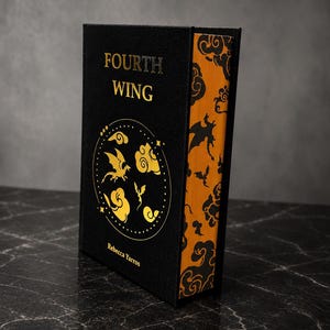 Peut inclure: Un livre relié noir intitulé Fourth Wing de Rebecca Yarros avec un lettrage doré et un motif circulaire représentant des dragons et des nuages sur la couverture. Les tranches des pages sont ornées d'un motif noir et orange de dragons.