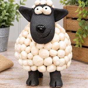 May include: A decorative sheep figurine featuring a matte black head with large white eyes and a glossy black nose. The body is covered in numerous small, off-white, rounded spheres representing wool, and the sheep stands on four black hooves.