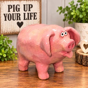 May include: A pink ceramic piggy bank with a glossy finish, featuring large floppy ears, a snout with two nostrils, and wide eyes. Behind the pig is a rectangular sign with a distressed white finish and the black text PIG UP YOUR LIFE.