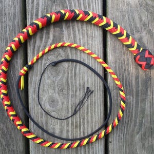 May include: A braided whip with a black, red, and yellow pattern. The whip is coiled on a wooden surface. The handle is black, and the main body of the whip is a combination of red, yellow, and black.