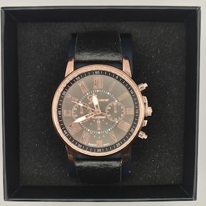 May include: A black leather strap watch with a rose gold bezel and a grey face. The watch features Roman numeral hour markers and three sub-dials. The watch is presented in a black box.